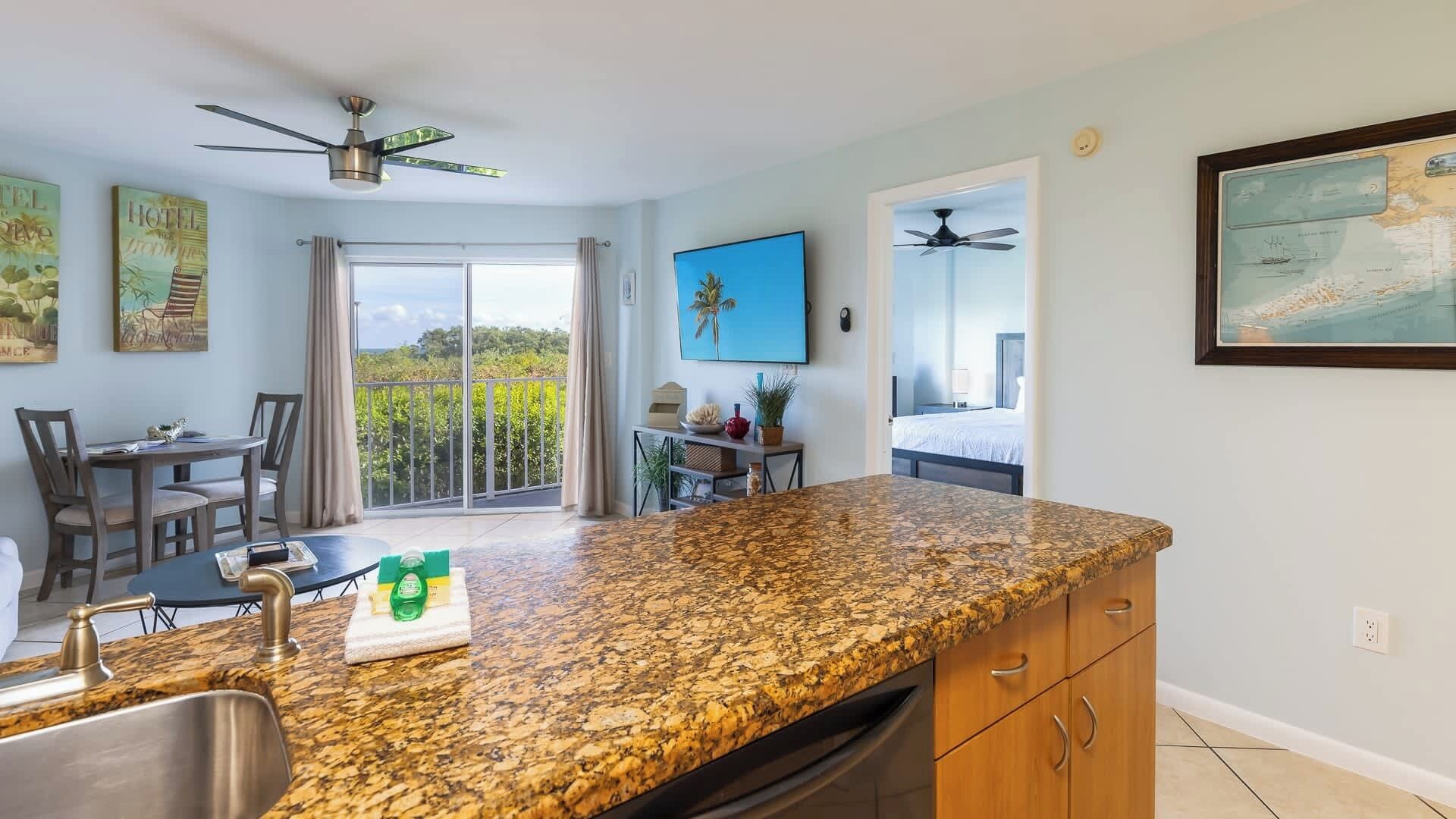 A kitchen with granite countertops, a dining area with a table and chairs, a balcony with a view, and a bedroom visible through an open door.