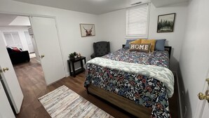 Un dormitorio con colcha estampada, almohada con la palabra 'HOME', una mesita de noche con una planta y un cuadro en la pared.