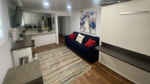 Una sala moderna con un sofá azul oscuro, una mesa de centro de madera y un tapete estampado. Se ve una cocina al fondo, con armarios blancos y electrodomésticos de acero inoxidable.