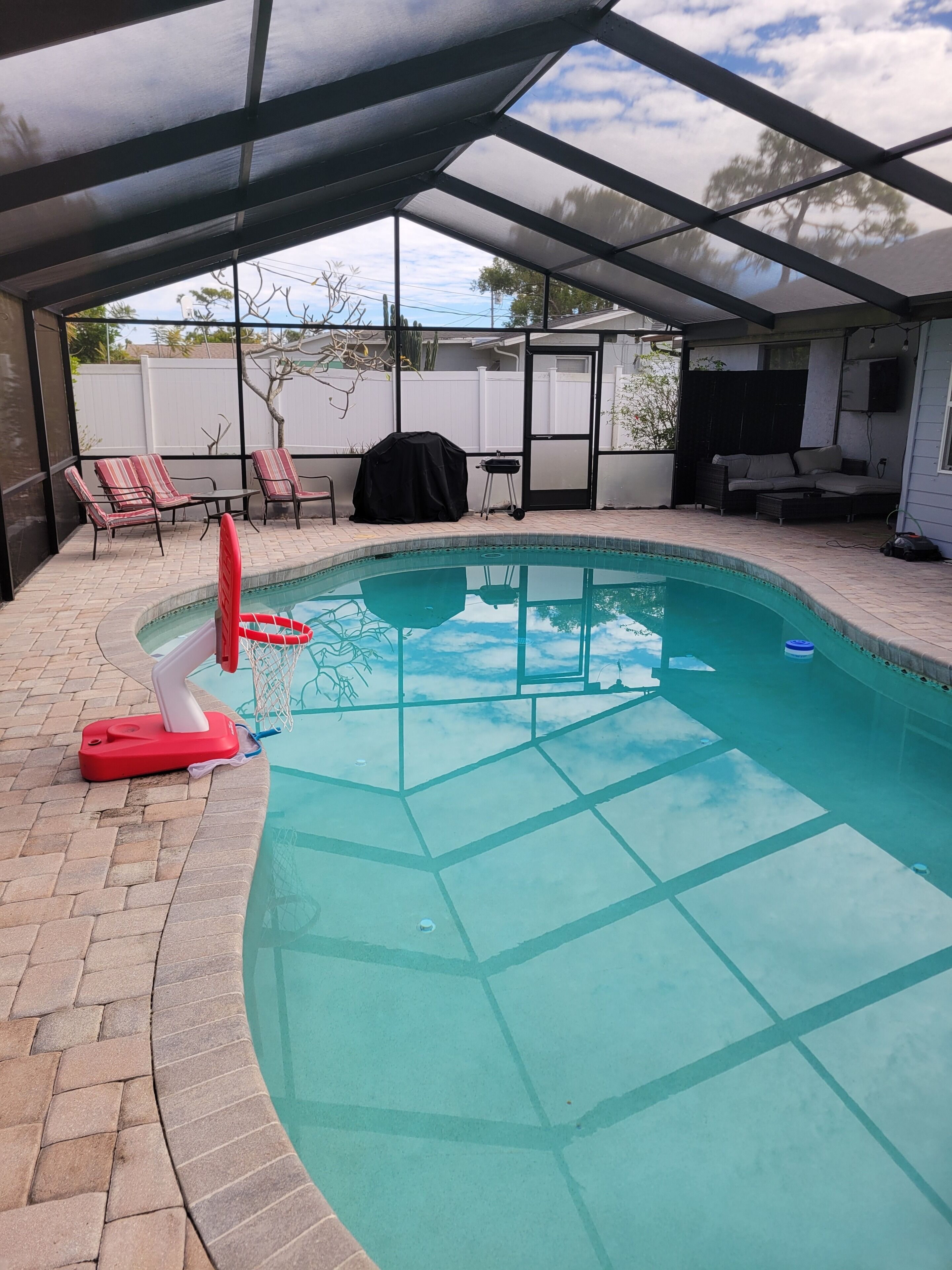 A swimming pool with a covered patio area, a basketball hoop, and outdoor seating.