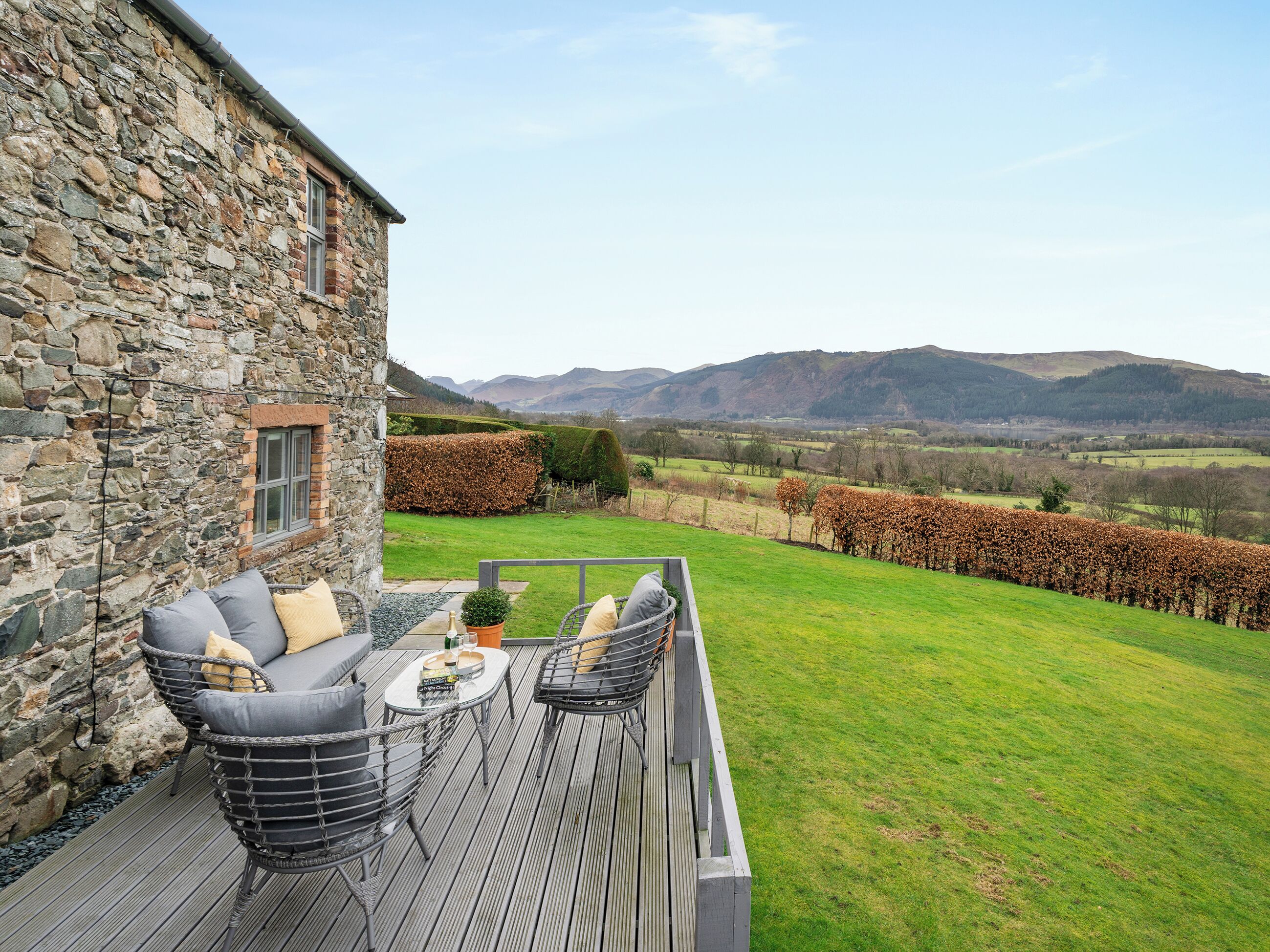 A stone building with a deck, outdoor seating, and a view of a grassy field and mountains.
