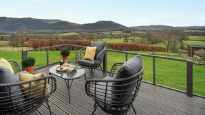 Una terraza con sillas de mimbre y una mesa, con vista a un paisaje rural con campos y colinas.
