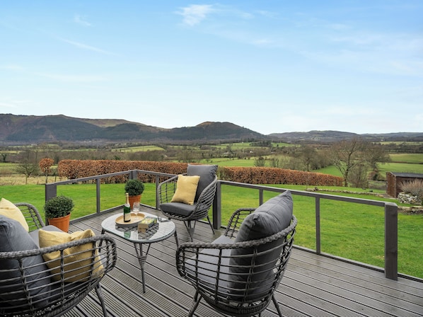 A deck with wicker chairs and a table, overlooking a rural landscape with fields and hills.