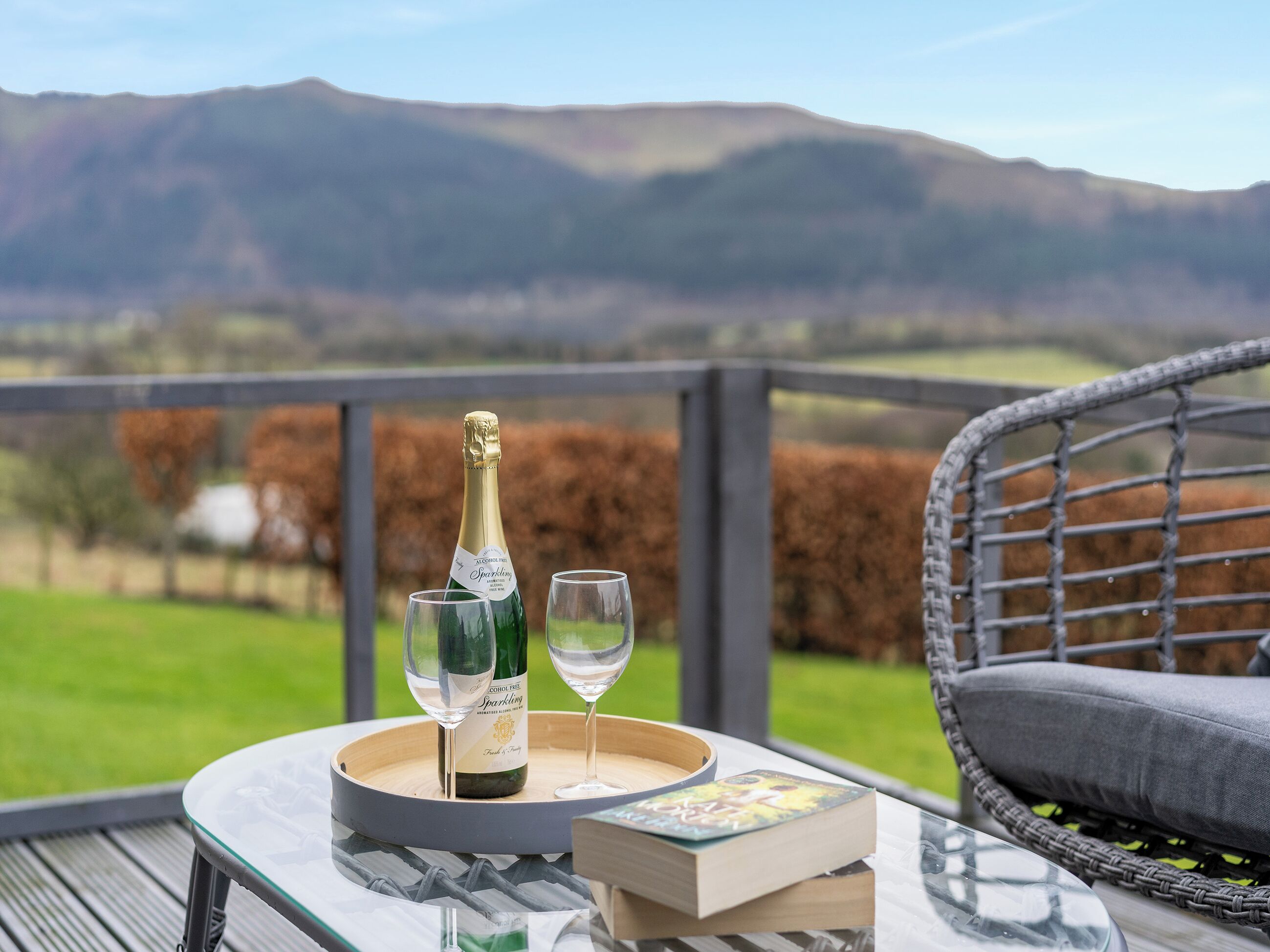 Et bord med champagneflaske, to glas og en bog på en balkon med udsigt over et smukt landskab.