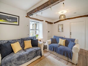 A living room with a grey sofa, a blue polka dot sofa, wooden beams, a hanging light fixture, and a window with a view of a stone building.