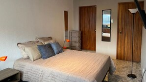 Een slaapkamer met een bed, kussens, een nachtkastje, een bureaulamp, een spiegel en een kledingkast.