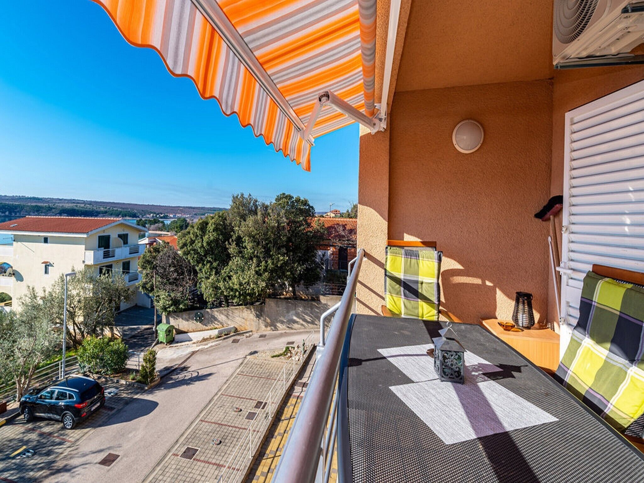 Un balcon donnant sur un parking et des immeubles, un coussin à carreaux et une petite lanterne.