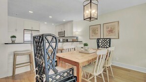 A modern kitchen with white cabinets, a microwave, and a dining area with a wooden table and white chairs. - 417 Seascape by Avantstay Beachfront Condo w/ Communal Pool Access (Isle of Palms)