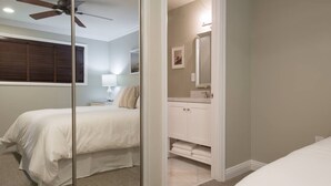 Et soveværelse med seng, natbord, lampe, ventilator i loftet, samt et badeværelse med vask, spejl og håndklædetørrestativ.