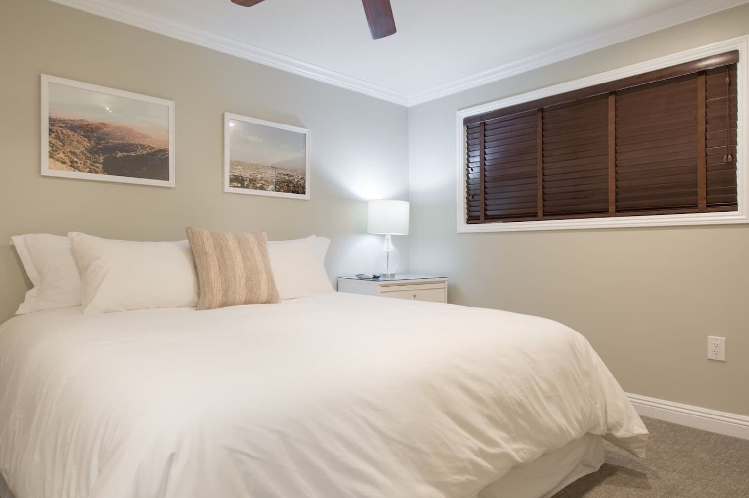 A bedroom with a large bed, two wall-mounted pictures, a bedside table with a lamp, and a window with blinds.