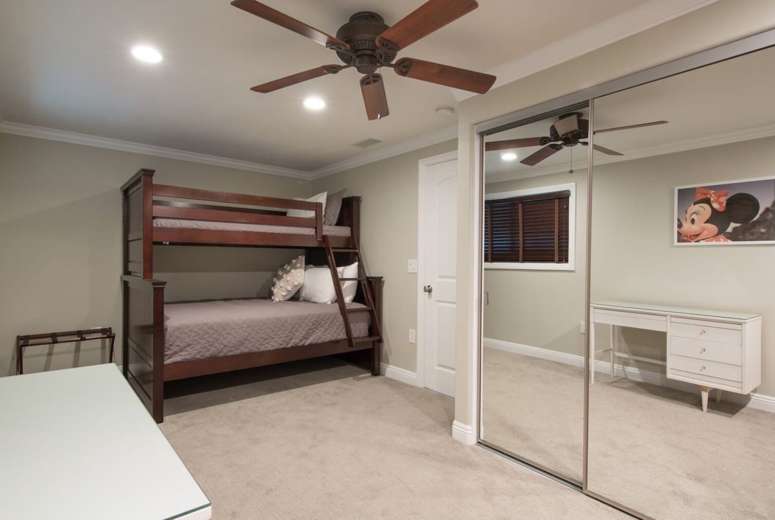 A bedroom with a bunk bed, a desk, a chair, a ceiling fan, and a door leading to another room.