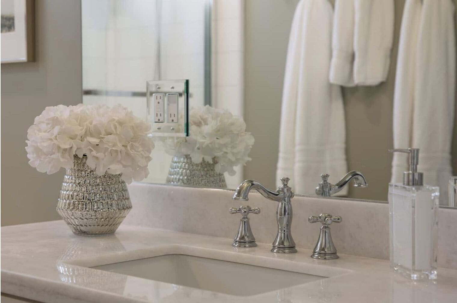 A bathroom with a white sink, two faucets, a mirror, and a vase with white flowers.