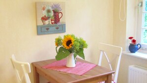 Un'area pranzo con tavolo in legno, due sedie bianche con cuscini a quadri bianchi e rossi, un vaso con girasoli, un quadro appeso al muro e una lampada a sospensione.