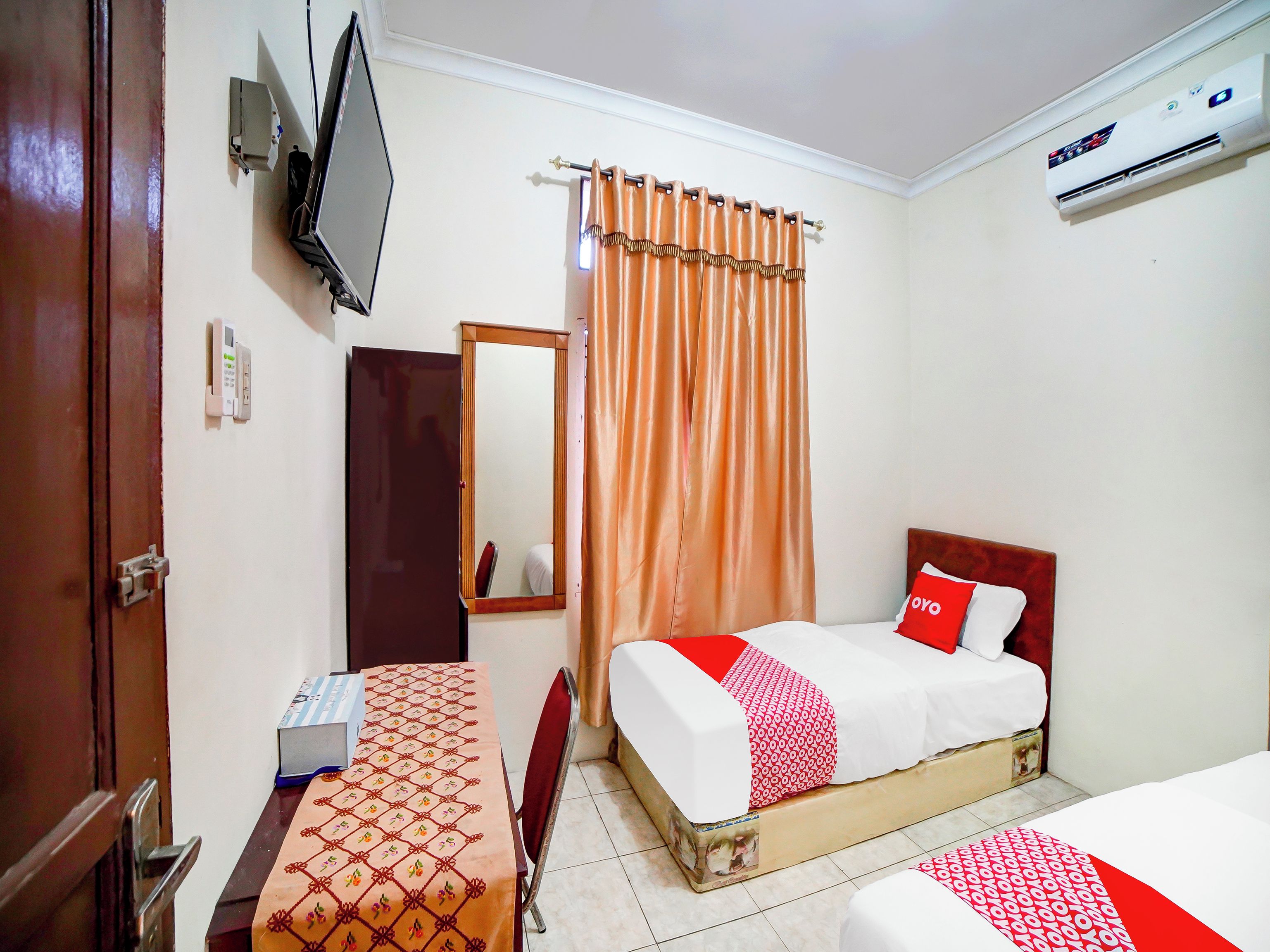 A hotel room with a bed, a bedside table, a mirror, a TV, and an air conditioning unit.