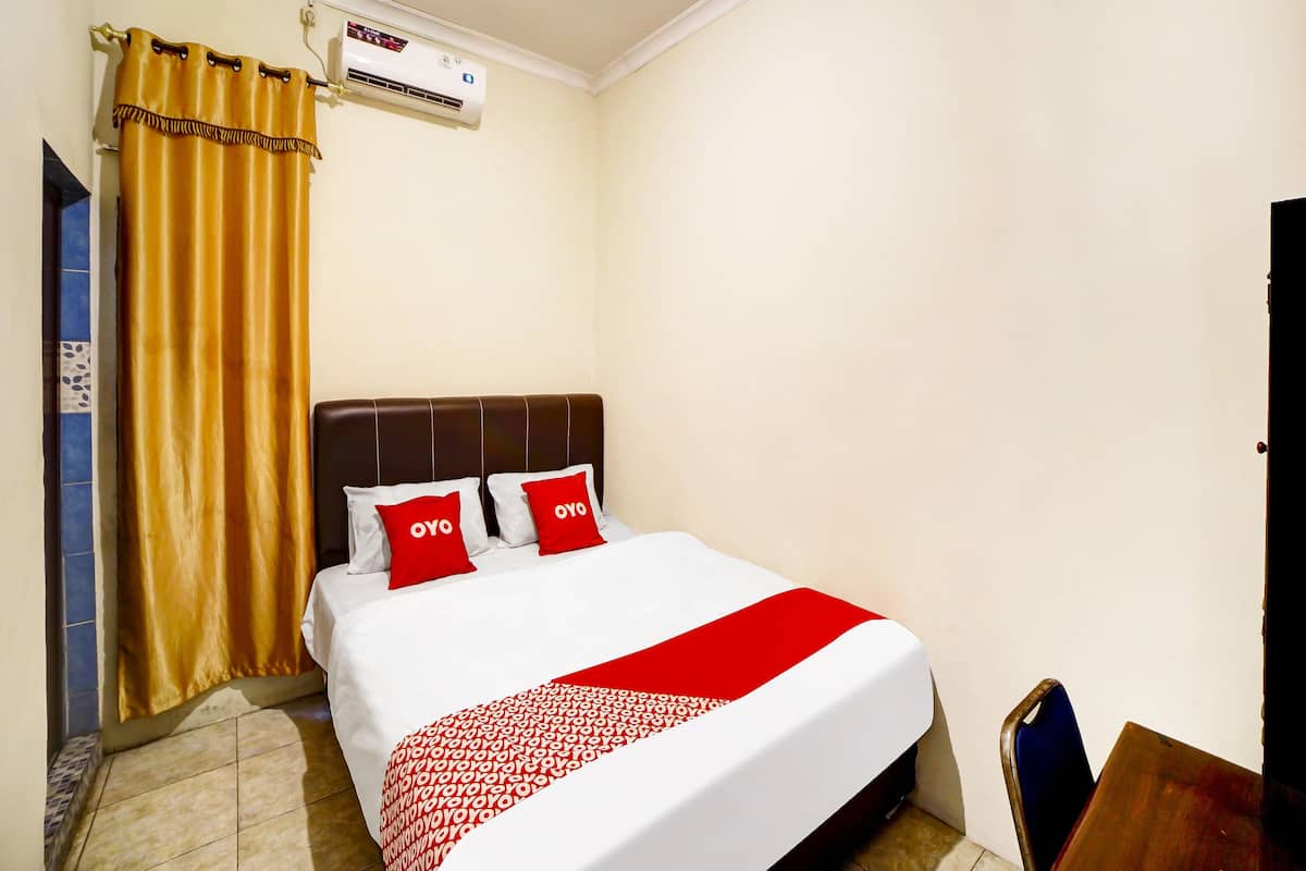 Ein Hotelzimmer mit einem Bett, zwei roten Kissen, einer weiß-roten Bettdecke, einem braunen Kopfteil, einem goldenen Vorhang und einer Klimaanlage an der Wand.