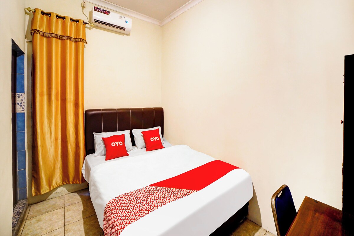 Habitación de hotel con una cama, dos almohadas rojas, una colcha blanca y roja, un cabecero marrón, una cortina dorada y un equipo de aire acondicionado en la pared.
