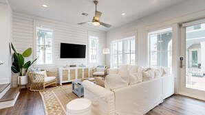 A spacious living room with a large white sofa, a ceiling fan, and a flat-screen TV mounted on the wall. - Serene newly built island home with outdoor pool, beach boardwalk & on-site golf (Port Aransas)