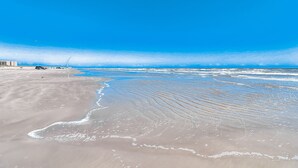 A beach with a clear sky, waves, and a few buildings in the background. - Serene newly built island home with outdoor pool, beach boardwalk & on-site golf (Port Aransas)