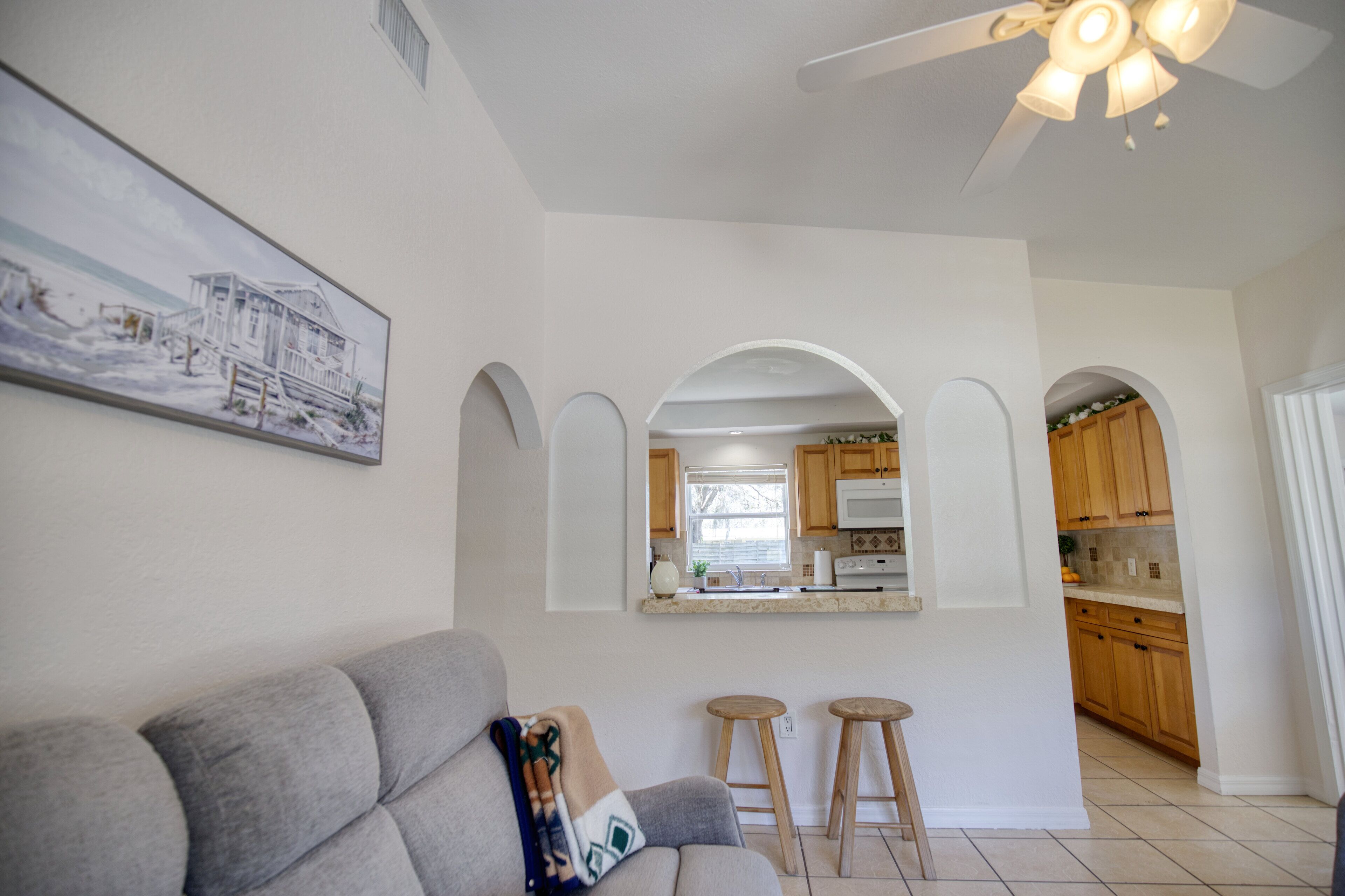 Un soggiorno con un divano grigio, due sgabelli di legno, una ventola a soffitto e una cucina visibile attraverso un arco.