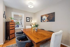 Un comedor con una mesa de madera, sillas tapizadas en gris y una silla blanca. Hay un aparador de madera con una planta encima, un cuadro enmarcado en la pared y un cuadro.