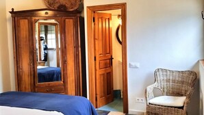 A room with a bed, a wicker chair, a wooden wardrobe with a mirror, and a door leading to another room.