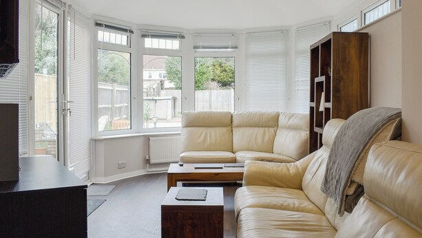 A living room with a beige sofa, a coffee table, and a TV stand. There are large windows with blinds, a radiator, and a wooden cabinet. - 2 bedroom accommodation in Poole (Poole)