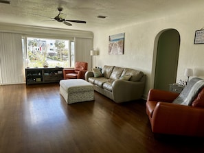 A living room with a sofa, ottoman, and a chair, a ceiling fan, and a view through a large window.