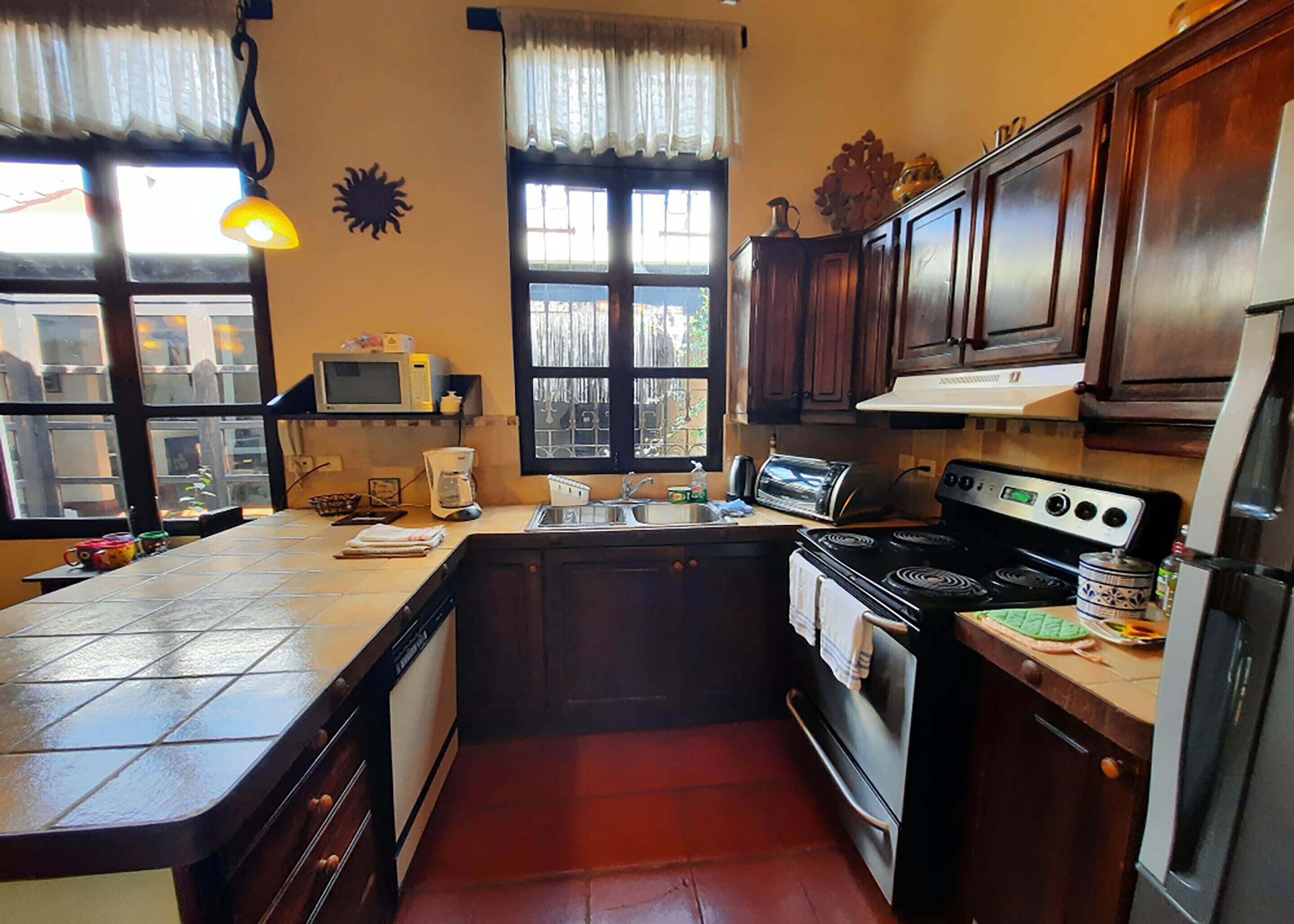 A compact kitchen with a stove, oven, microwave, and sink. There is a window with curtains, a hanging light, and a small shelf with a microwave and a coffee maker.
