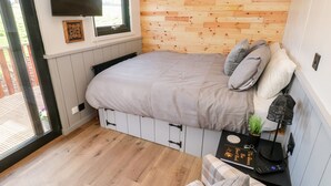 A cozy bedroom with a bed, a window, a wall-mounted TV, and a small table with a plant and a speaker. - The Queen Bee (Pembroke)
