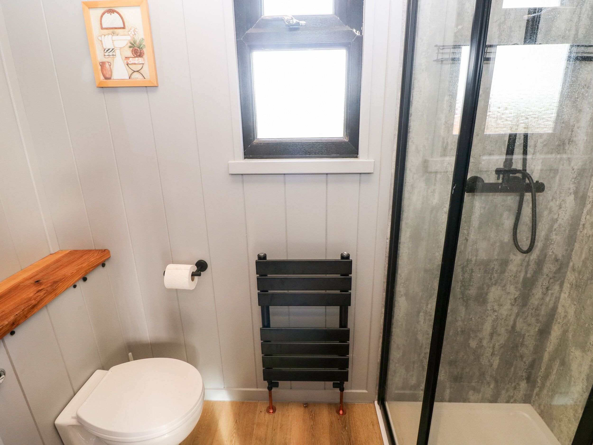 A bathroom with a toilet, a wooden shelf, a towel rack, a window, and a glass shower enclosure.