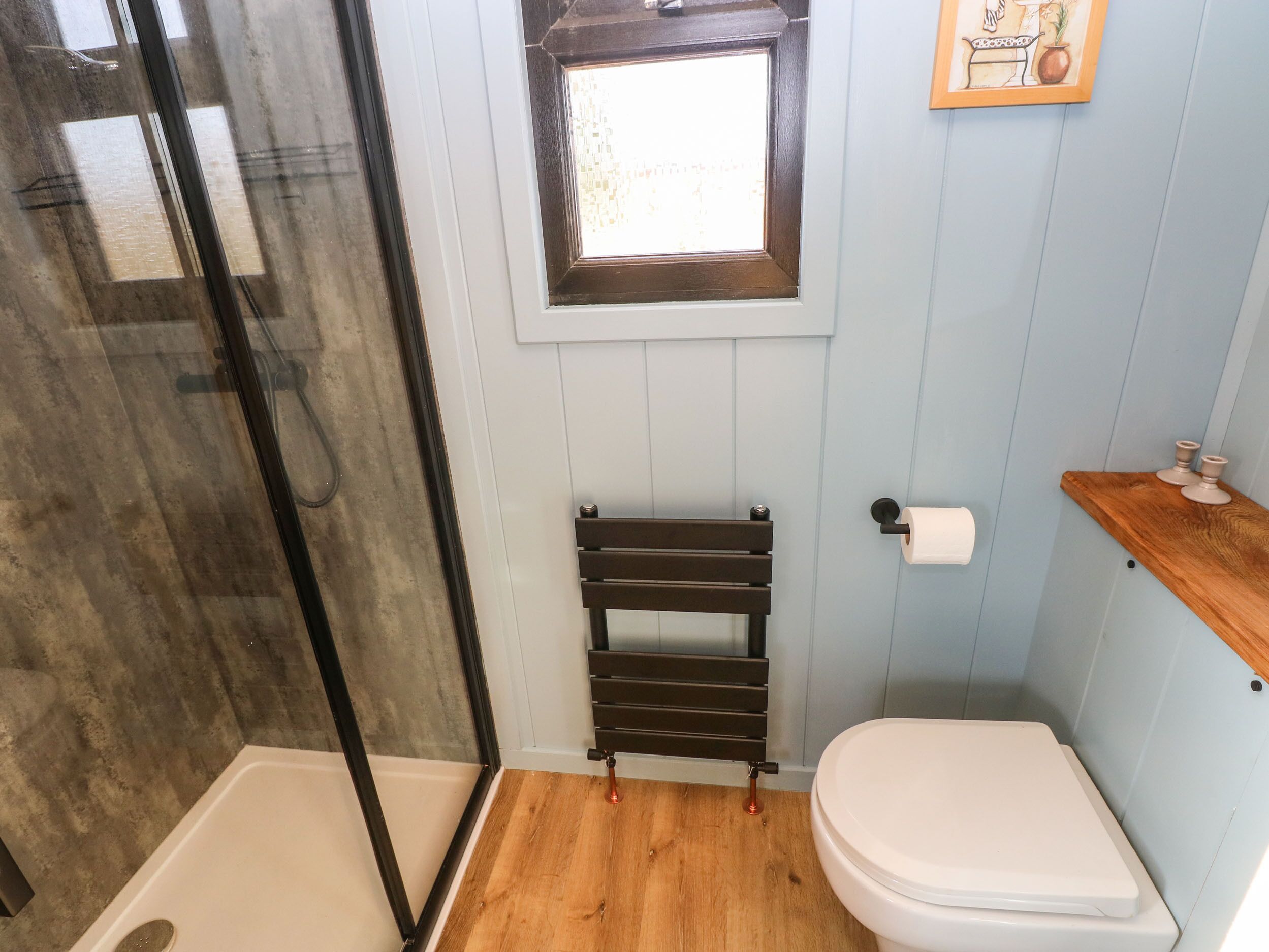 Un baño con cabina de ducha de vidrio, inodoro blanco, suelo de madera, un ventanal pequeño y un perchero para toallas.