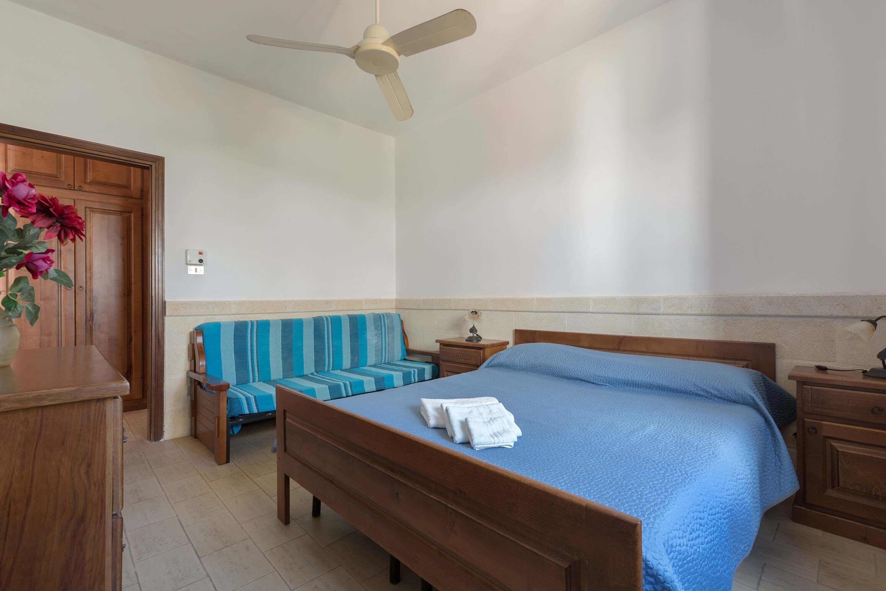 Ein Schlafzimmer mit einem hölzernen Bett, einem blau gestreiften Sofa, einem Nachttisch aus Holz und einem Deckenventilator.