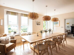 A spacious dining area with a wooden table and chairs, large windows with curtains, and hanging pendant lights. - Treveddoe Farmhouse (Bodmin)