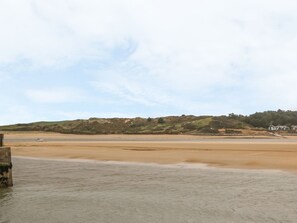 A coastal landscape with a sandy beach, a stone structure in the foreground, and a hilly background with buildings. - Treveddoe Farmhouse (Bodmin)