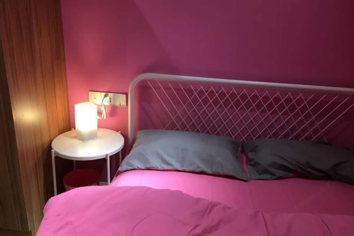 A bed with a metal frame, a round bedside table with a lamp, and a red stool.
