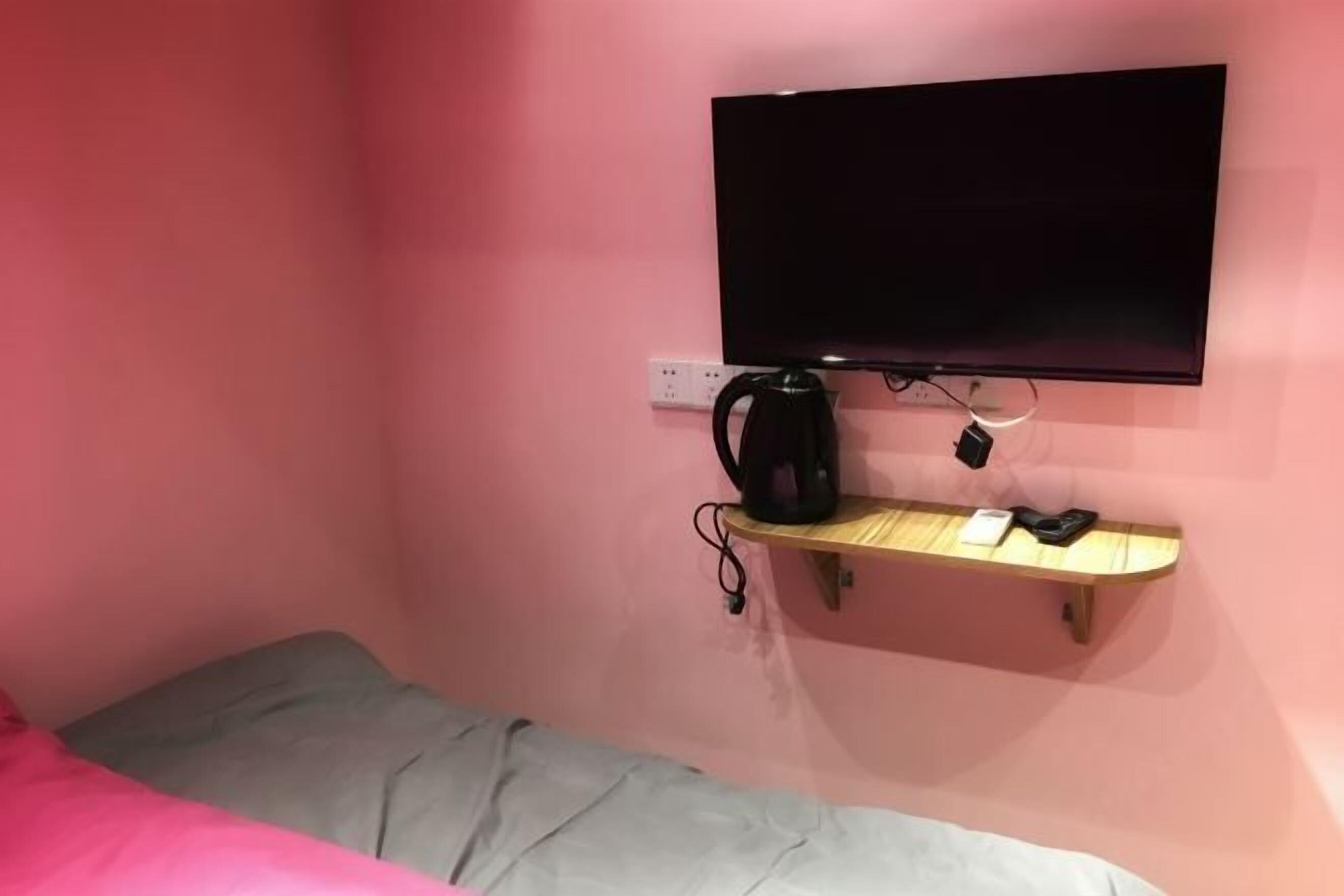 A small room with a bed, a television mounted on the wall, and a shelf with a kettle and a phone.