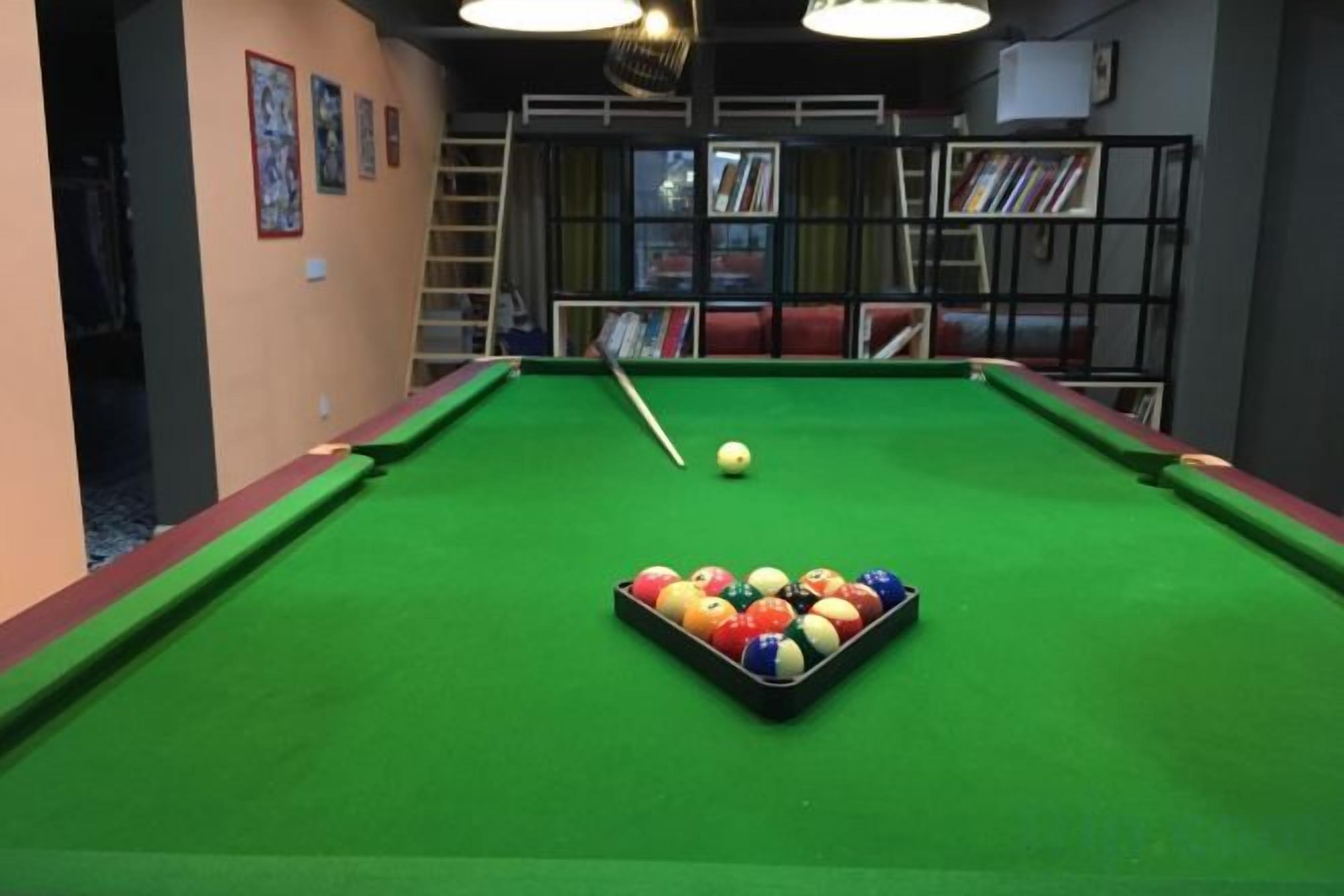 A pool table with balls, a cue stick, and a bookshelf in the background.