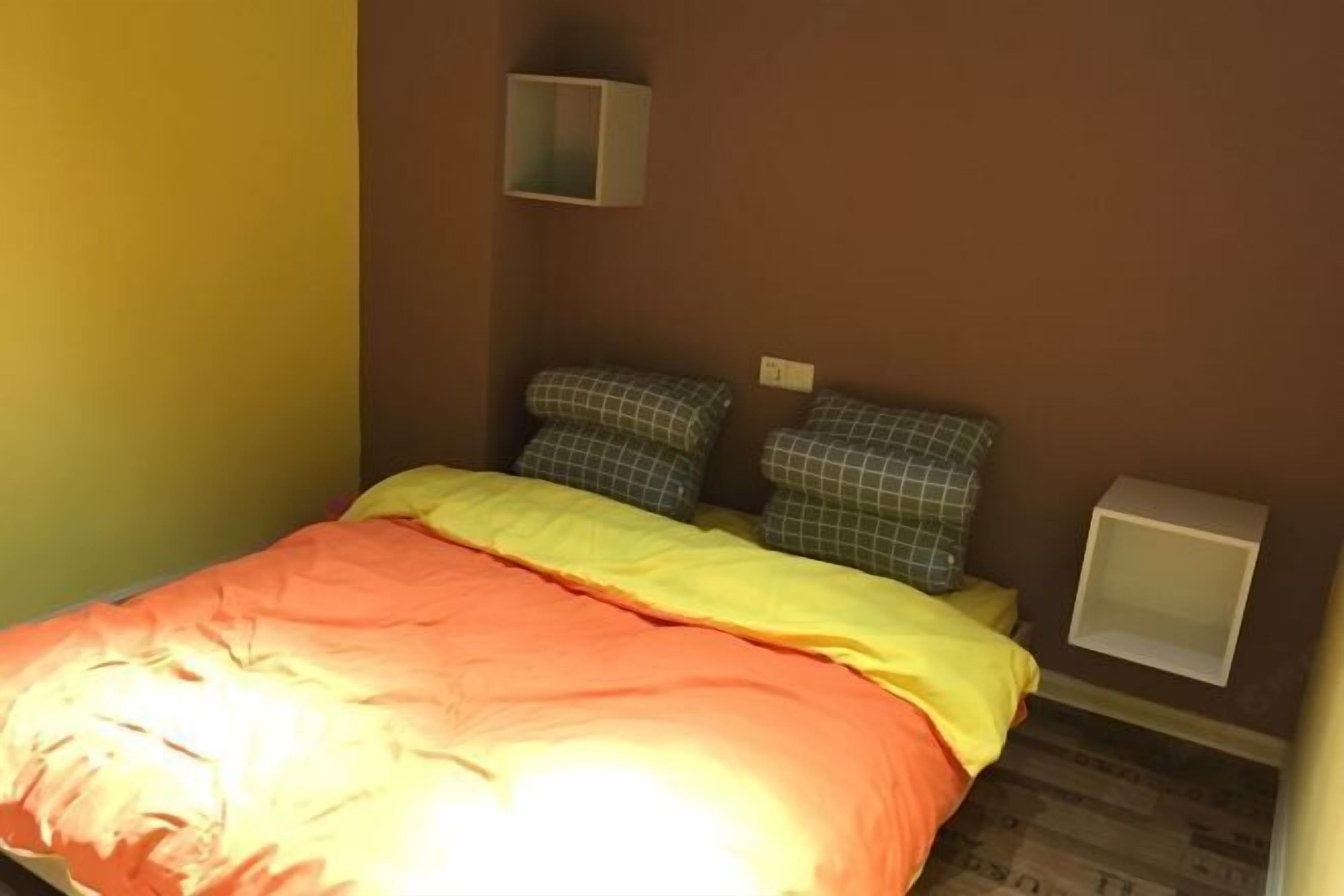Quarto pequeno com uma cama de solteiro, coberta por um edredom amarelo e laranja, piso de madeira e uma parede lisa com uma pequena prateleira.