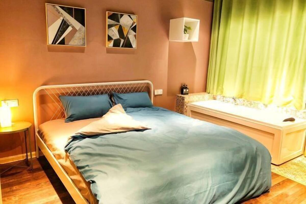 A bedroom with a bed, a bedside table, a wall-mounted lamp, and a green curtain.