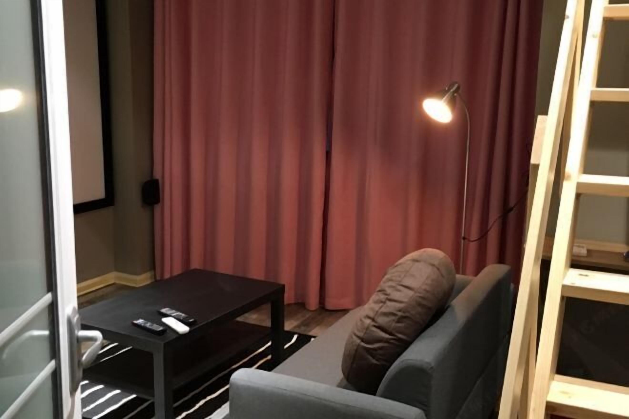 A room with a grey sofa, a small black table with remotes, a lamp, and a wooden ladder.