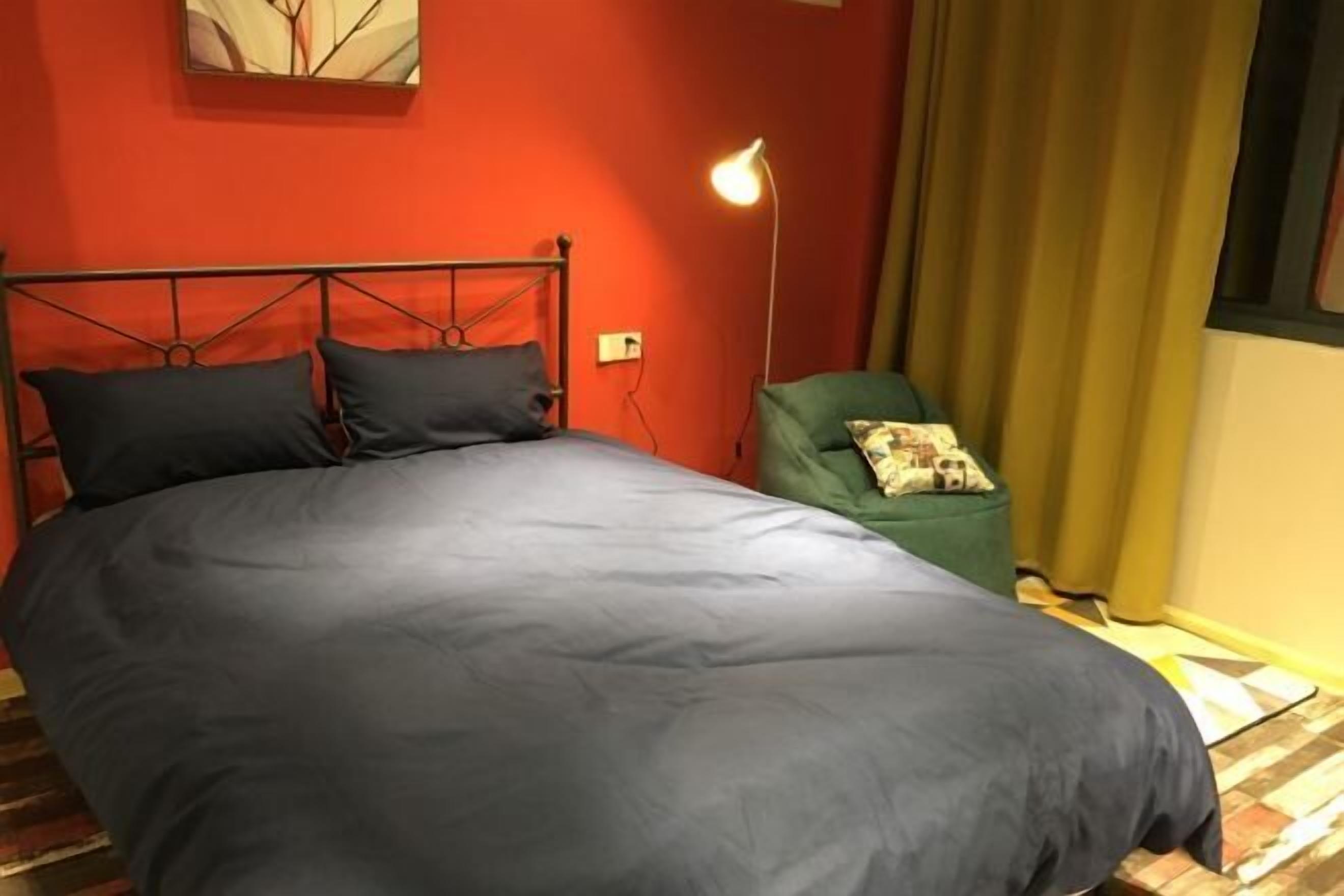 Een bed met metalen frame, een groene fauteuil, een vloerlamp en een raam met gordijnen.