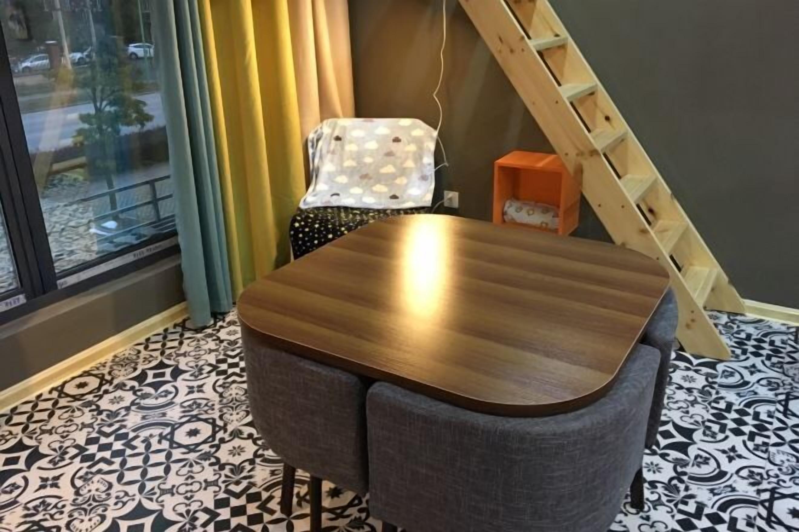 A room with a wooden table and two grey chairs, a window with curtains, a patterned floor, and a wooden ladder against the wall.