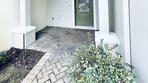 Een overdekt buitengebied met een deur, een kleine plant en een pad van bakstenen.