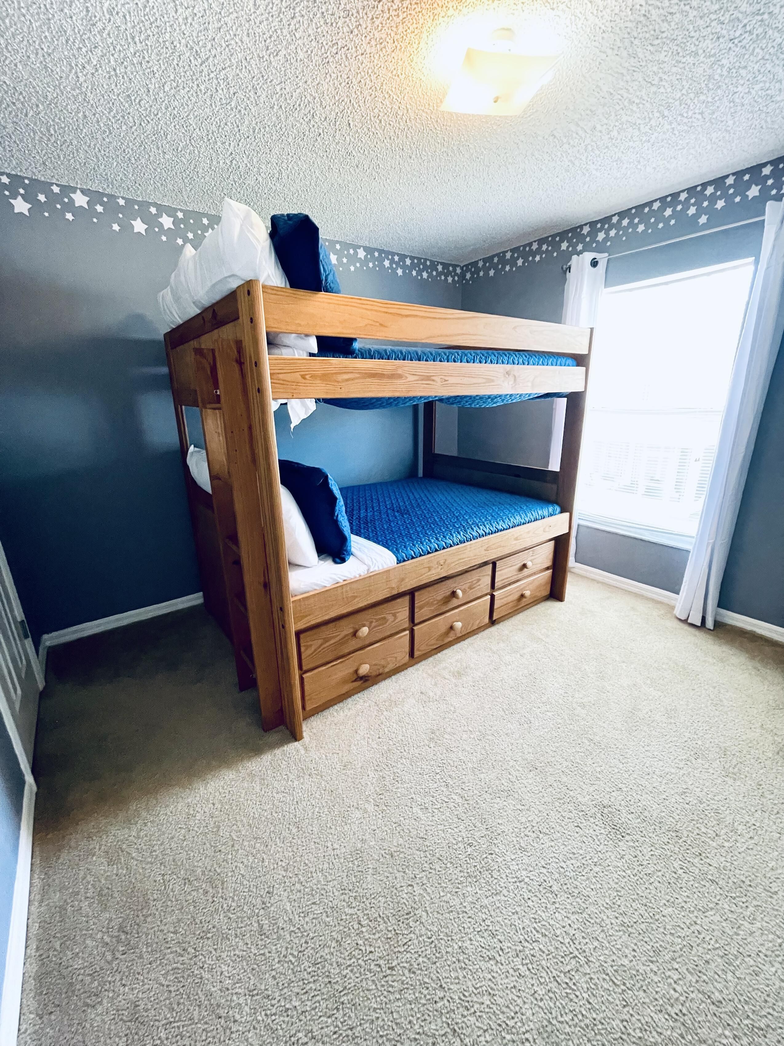 A bunk bed with a wooden frame and blue bedding, a carpeted floor, and a window with curtains.