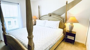 Een hemelbed met houten hoofdeinde, nachtkastje met een lamp en uitzicht buiten door een raam met blinds.