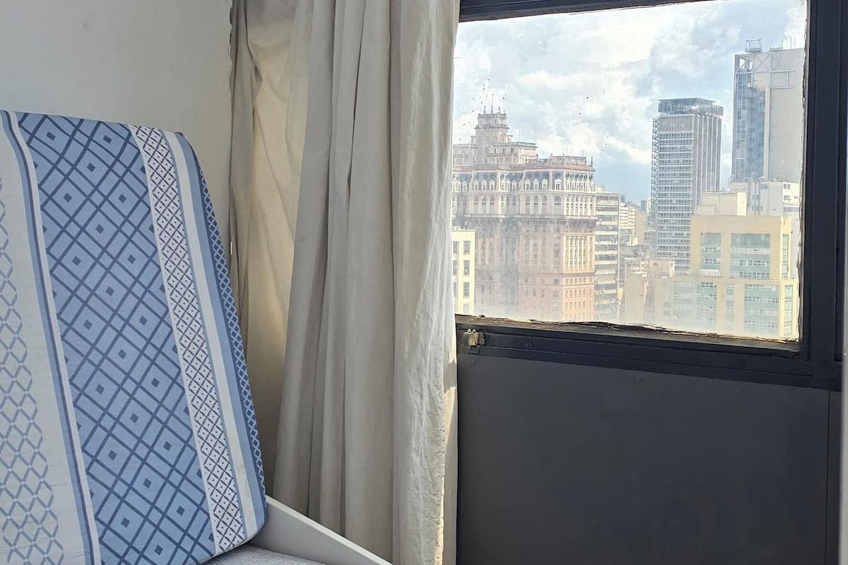 Una habitación pequeña con cama, ventana con vista a la ciudad y ventilador de techo.
