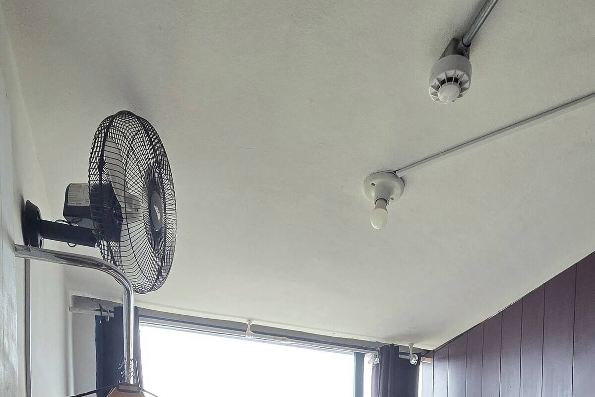 Una habitación con una cama individual superpuesta, un ventilador de pie, una ventana con cortinas y un suelo de madera con motivo de espiga.