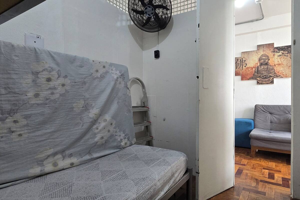Una habitación pequeña con una cama litera, un ventilador de pie, un estante y un cuadro de Buda colgado en la pared.