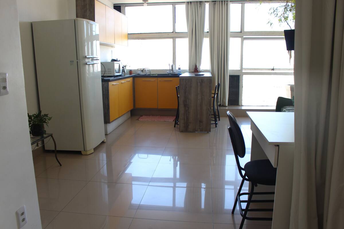 Cocina compacta con refrigerador, gabinetes amarillos, una isla de madera y un área de comedor con sillas.