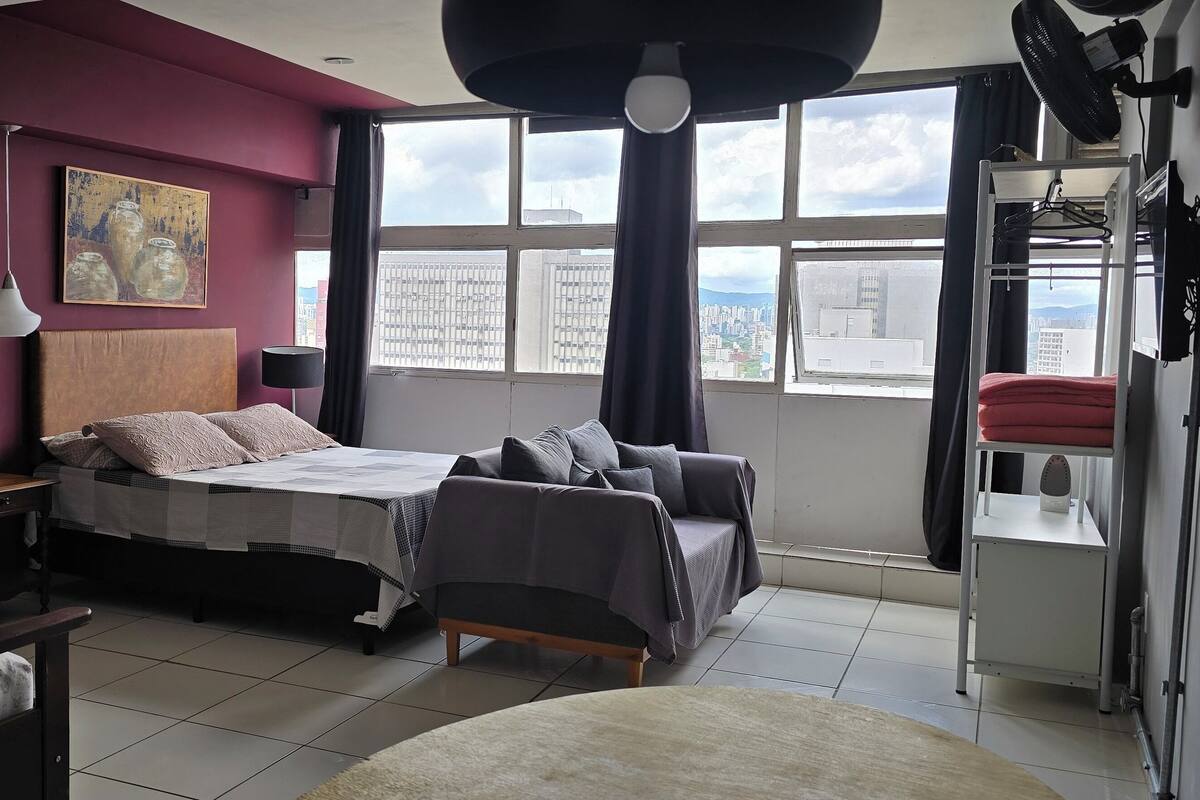 Un dormitorio moderno con cama, sofá, escritorio y un amplio ventanal con vista a la ciudad.